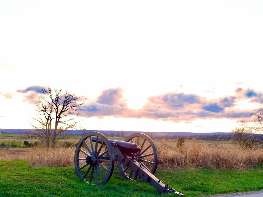 A historic cannon sits on a grassy hill overlooking a field with a bare tree under a cloudy sunset sky.