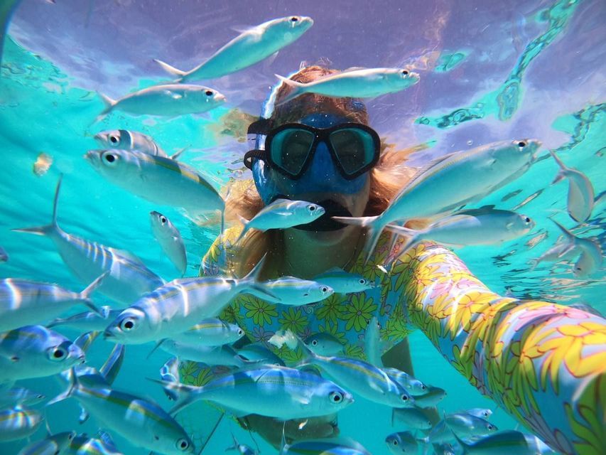 Una persona con maschera da snorkeling e muta colorata si fa un selfie nuotando sott'acqua, circondata da un banco di pesci.