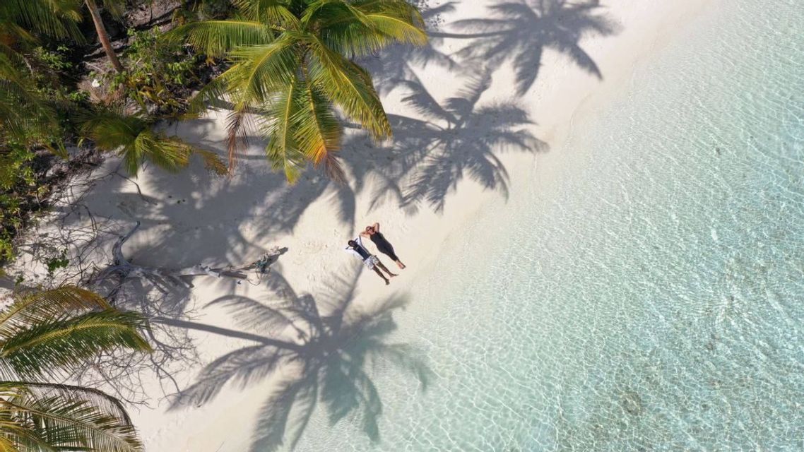 Una vista aerea di due persone che si rilassano su una spiaggia di sabbia bianca, con acqua cristallina e ombre di palme.