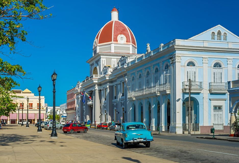 A vintage blue car drives on a street next to a large building with a red dome under a clear blue sky.