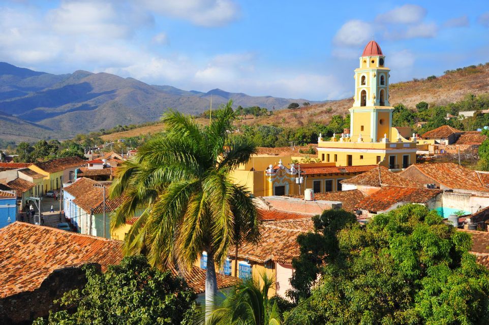 Un campanile giallo si erge su una città con tetti in terracotta, con palme e montagne verdi sullo sfondo.