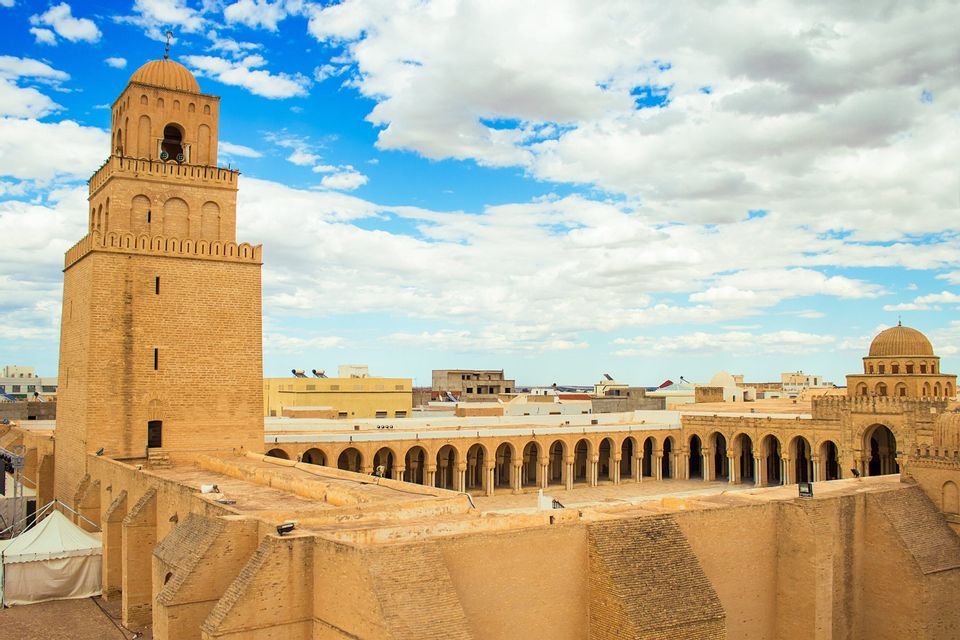 Una veduta dall'alto di una storica moschea in arenaria, con un minareto prominente e un cortile ad arco, sotto un cielo blu con nuvole.