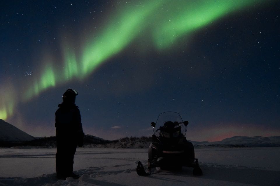 Una persona junto a una moto de nieve en un paisaje nevado nocturno, contemplando la aurora boreal verde que llena el cielo estrellado.