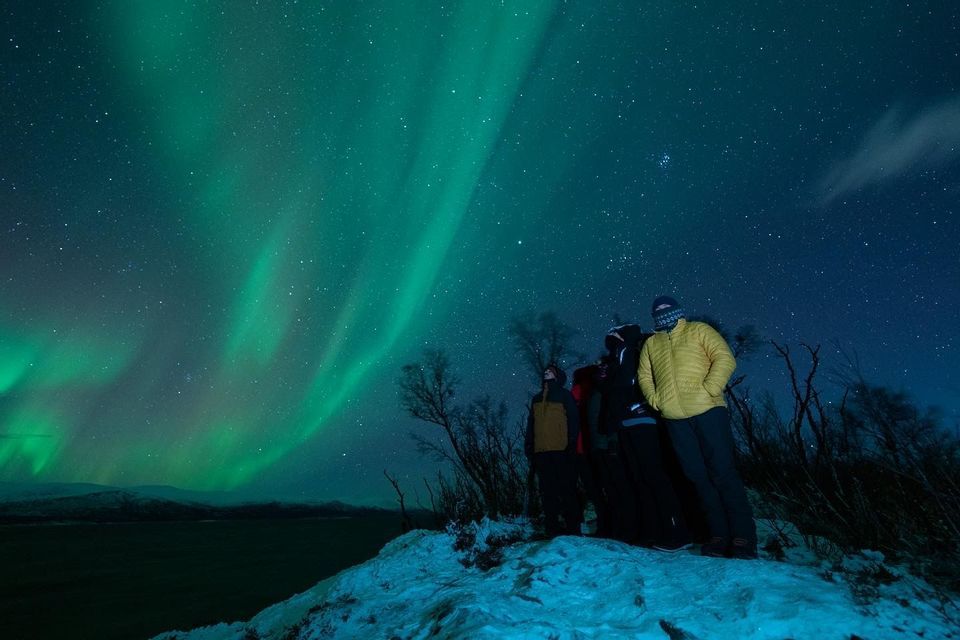 Un grupo de WeRoad en una colina nevada por la noche, contemplando la aurora boreal verde bajo un cielo estrellado.