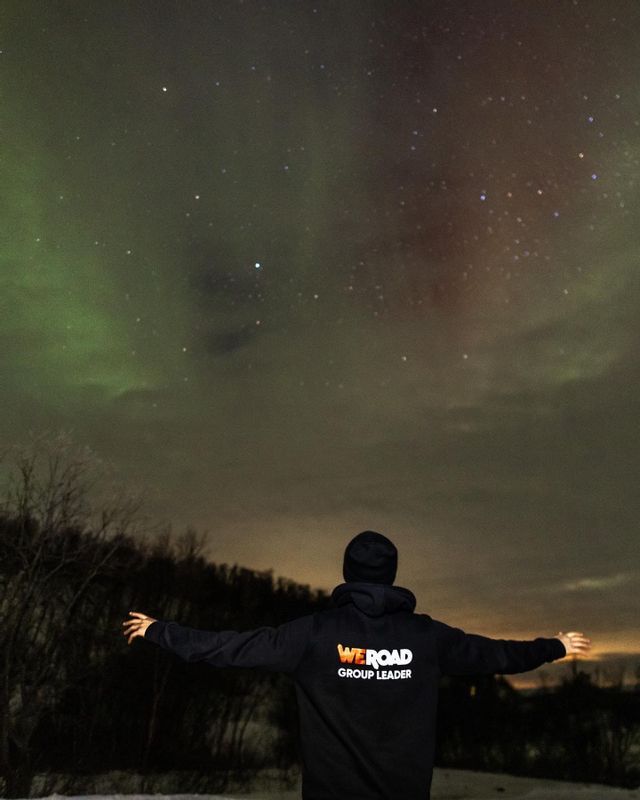 A WeRoad Group Leader stands with arms outstretched, looking up at the aurora borealis and starry night sky.