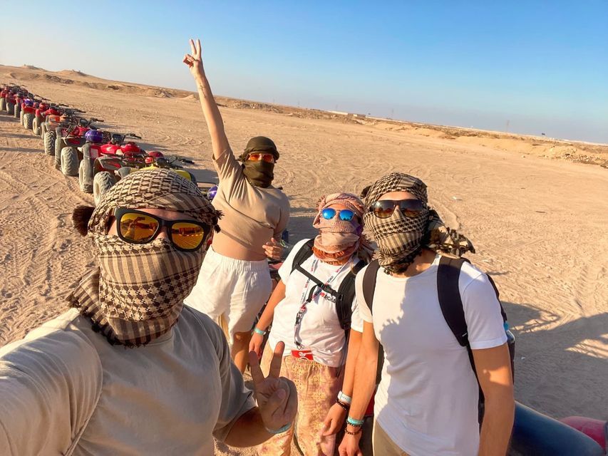 Un viaje en grupo de WeRoad de cuatro personas con kefias se toma una selfie en el desierto junto a una fila de quads.