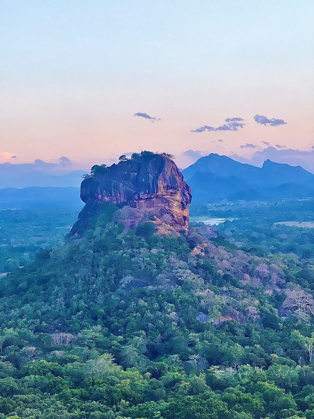Una grande formazione rocciosa alberata si erge su una fitta giungla verde, con montagne distanti sotto un cielo dai colori pastello al crepuscolo.