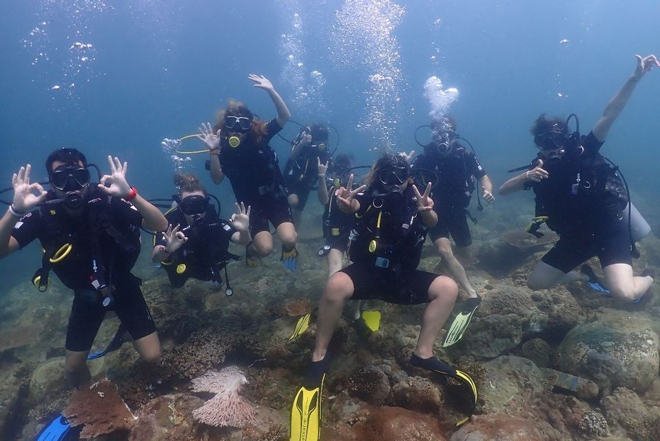 Un gruppo WeRoad in attrezzatura subacquea posa per una foto sottomarina, facendo segni con le mani sopra una scogliera rocciosa.