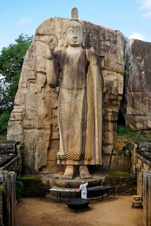 Una persona vestita di bianco si trova ai piedi di una colossale statua di Buddha scolpita nella roccia, vista dall'alto.