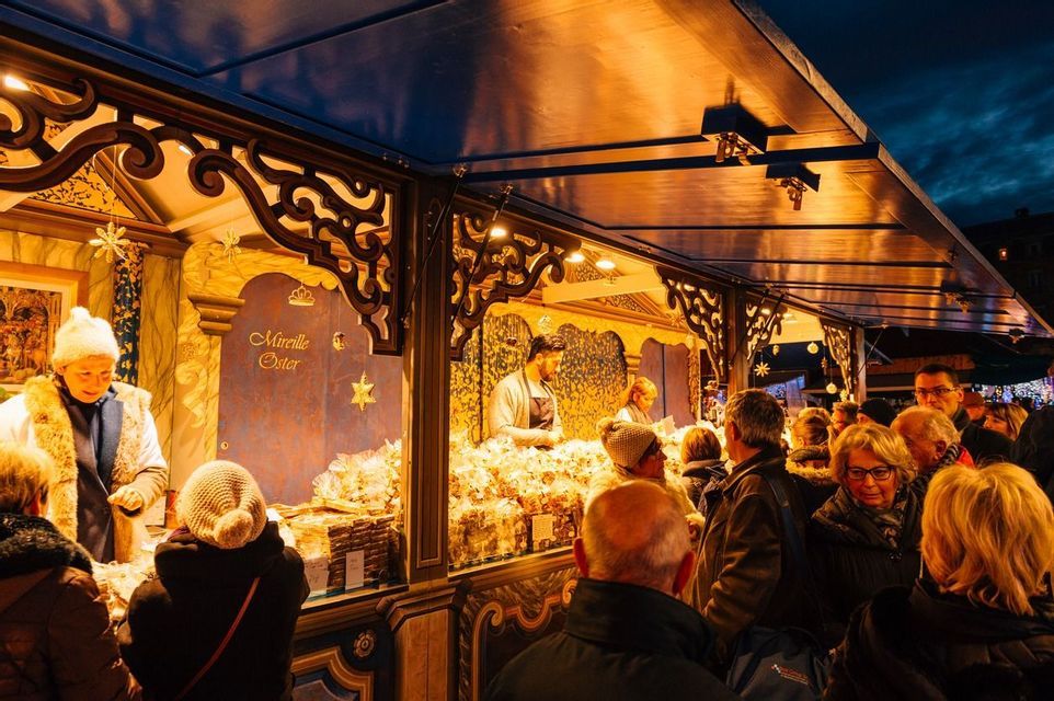 A crowd of people shops at a brightly lit, ornate wooden stall in an outdoor market at dusk.