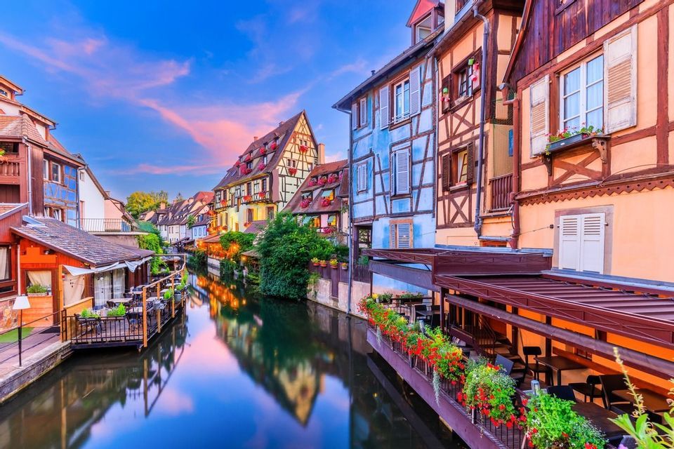 Colorful half-timbered buildings with balconies full of flowers line a calm canal, reflecting in the water at sunset.