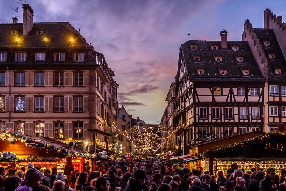 A crowded Christmas market fills a narrow street between old buildings, illuminated by festive lights under a purple sky.