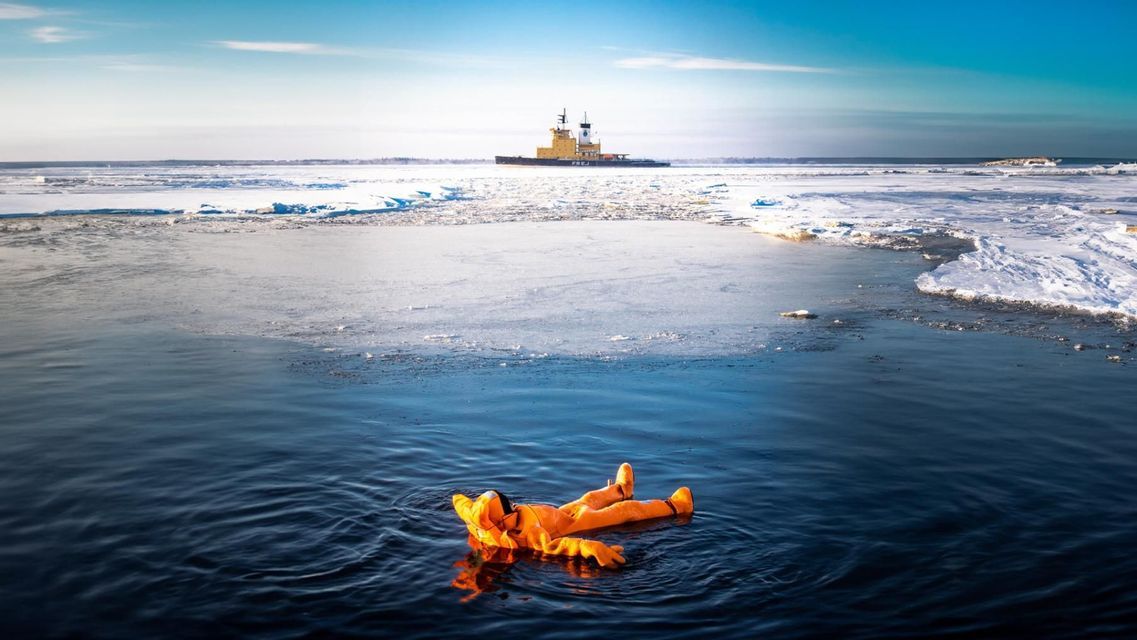 Une personne en combinaison de survie orange flotte sur le dos dans l'eau glacée, avec un brise-glace au loin.