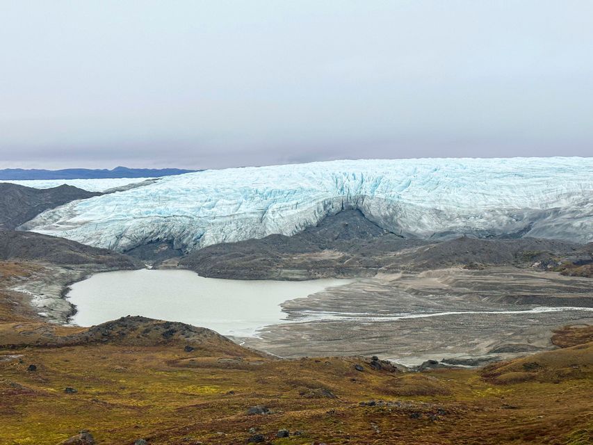 Una vista panoramica di un imponente ghiacciaio dietro un lago glaciale lattiginoso, con terreno roccioso e tundra marrone in primo piano.