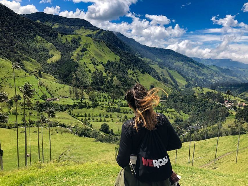 Una mujer con una mochila WeRoad contempla un exuberante valle verde con colinas ondulantes y palmeras altas bajo un cielo parcialmente nublado.