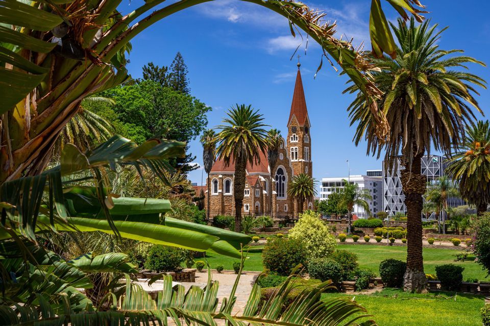 Una chiesa in pietra con una guglia rossa si erge in un parco curato incorniciato da palme tropicali sotto un cielo azzurro e limpido.