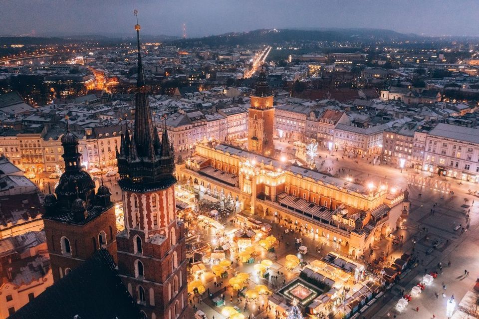 Una vista aerea di una vivace piazza cittadina di notte, con un mercatino di Natale illuminato e neve sui tetti.
