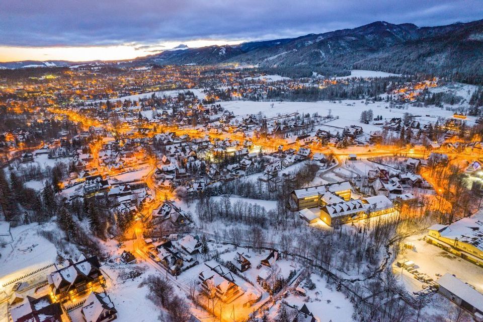Eine Luftaufnahme einer schneebedeckten, in der Abenddämmerung von Lichtern erleuchteten Stadt mit einer Bergkette im Hintergrund.