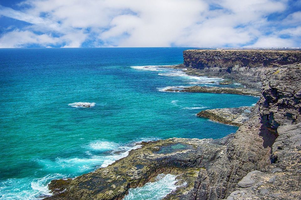 Una costa frastagliata e rocciosa costeggia un oceano turchese con piccole onde bianche sotto un cielo azzurro con nuvole soffici.