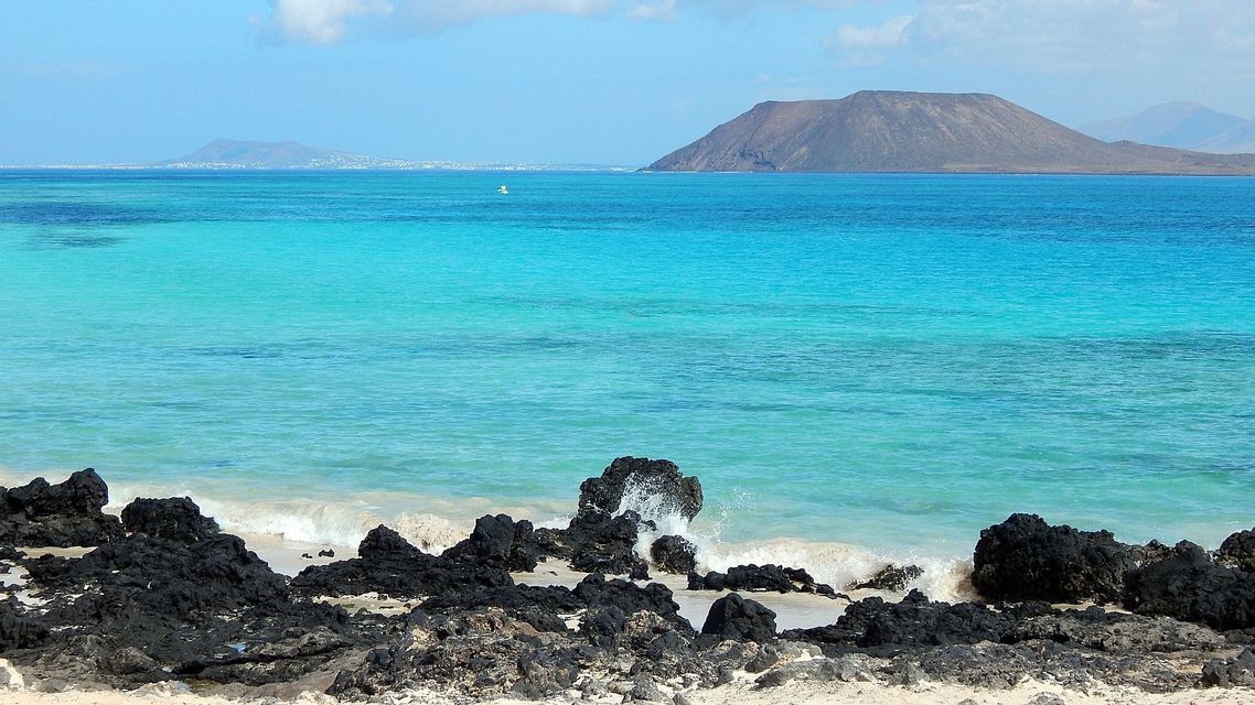 Agua turquesa baña rocas volcánicas negras en una playa de arena, con una isla volcánica en la distancia bajo un cielo azul.