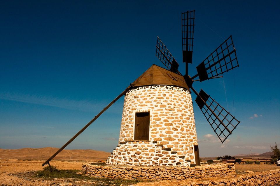 Un molino de viento tradicional de piedra blanca con aspas de madera se alza en un paisaje seco y árido bajo un cielo azul claro.
