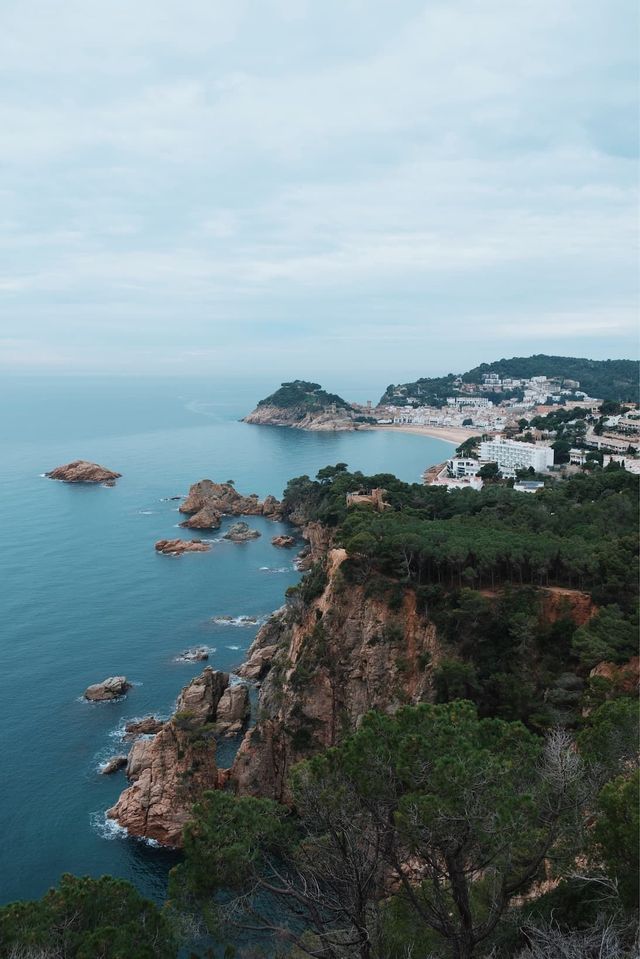 A high-angle view of a rocky, tree-covered coastline leading to a seaside town with a sandy beach under a cloudy sky.