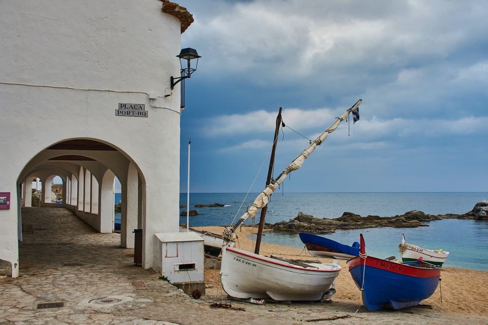 Fishing boats rest on a sandy beach next to a white building with an arched walkway, overlooking the sea under a cloudy sky.