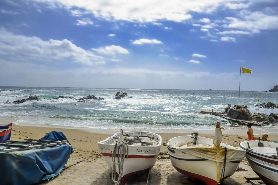 Small fishing boats sit on a sandy beach with a choppy sea and a cloudy sky in the background.