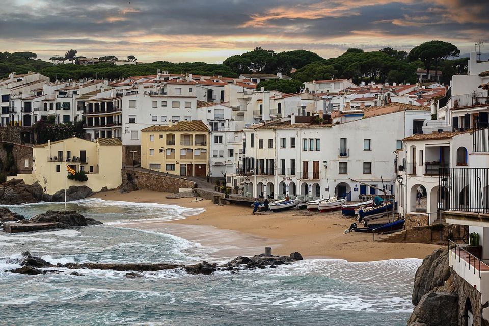 White buildings of a seaside village line a sandy cove with boats on the shore and waves breaking on the rocks.