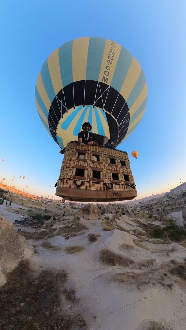 Selfie grand-angle d'une personne en montgolfière survolant un paysage rocheux au lever du soleil, avec des dizaines d'autres ballons dans le ciel.