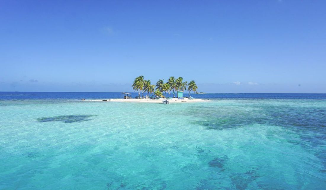 Una piccola isola sabbiosa con palme e una capanna si erge in acque limpide e turchesi sotto un cielo blu.