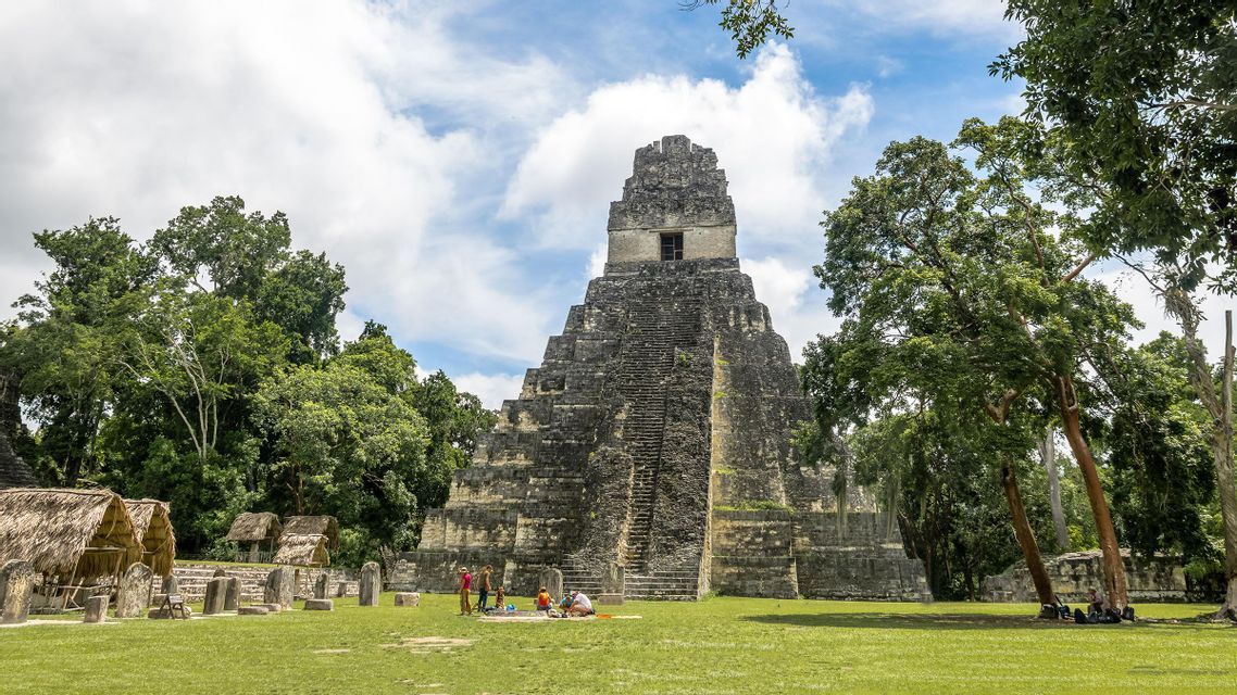 Una grande piramide a gradoni di pietra si erge in una radura erbosa circondata dalla giungla, con alcune persone che riposano alla sua base.