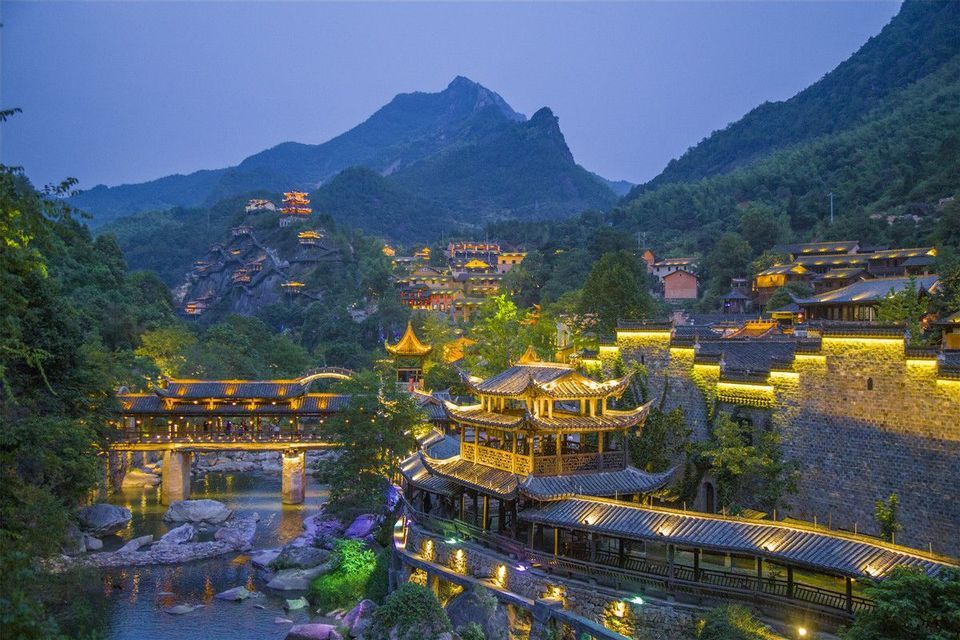 Un villaggio illuminato con architettura tradizionale e un ponte su un fiume, incastonato in una verde valle di montagna al tramonto.