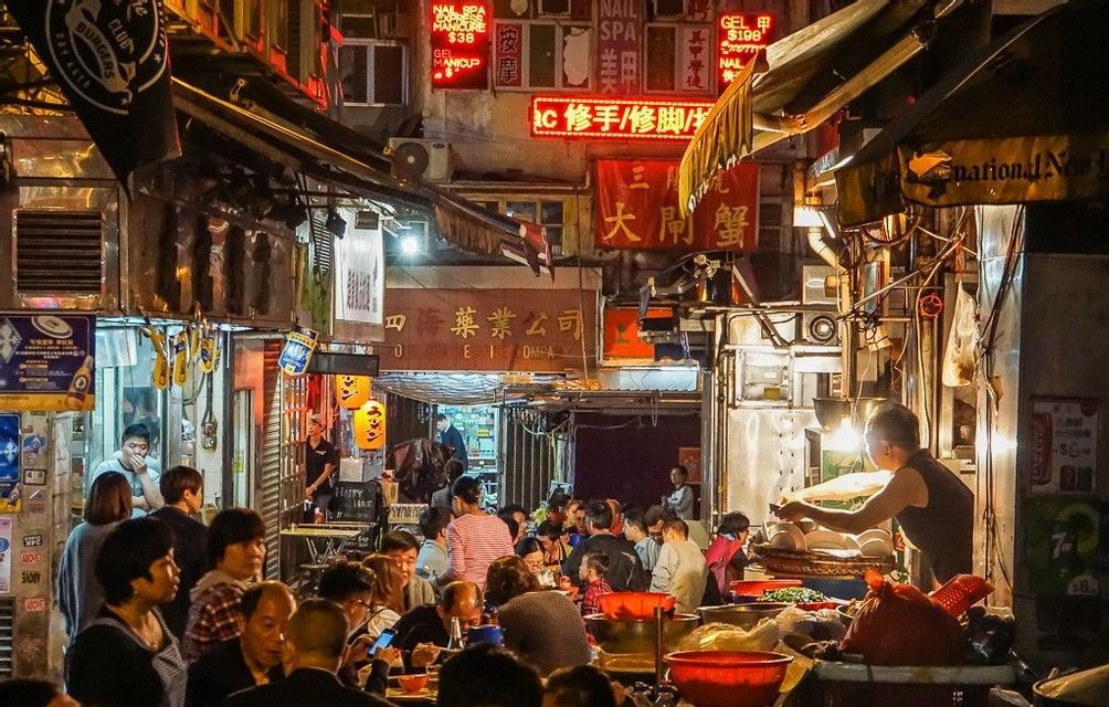 Una grande folla di persone che mangiano ai tavoli all'aperto in un affollato vicolo cittadino di notte, illuminato da insegne al neon e bancarelle di cibo.