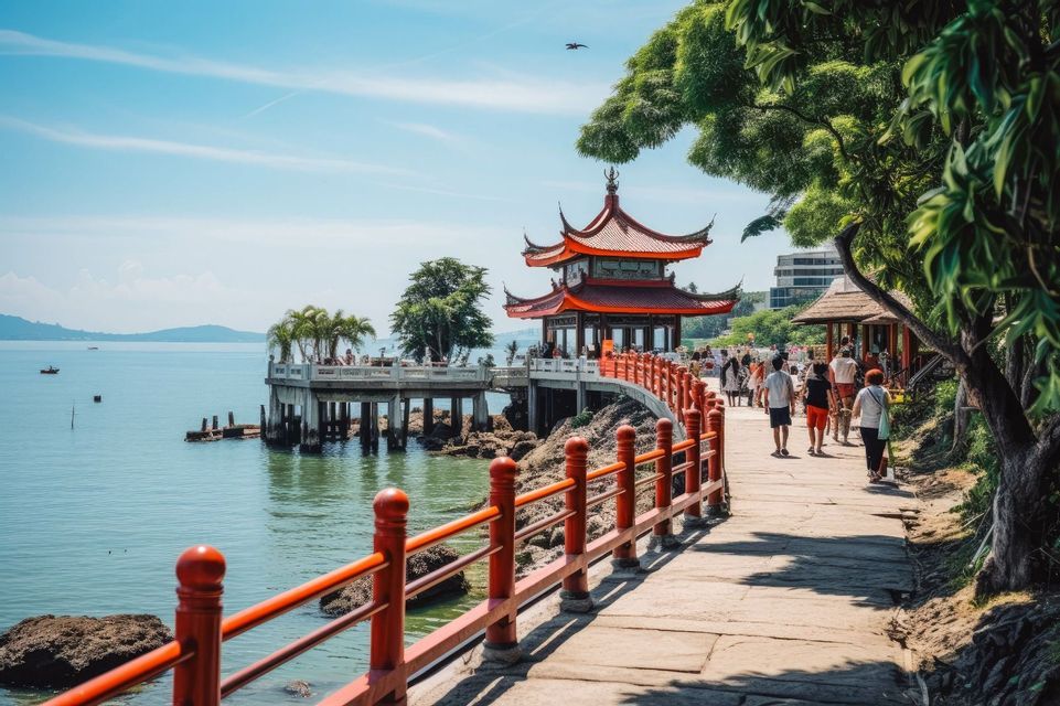 La gente camina por un sendero costero con una barandilla roja hacia una pagoda tradicional construida en un muelle sobre el agua.