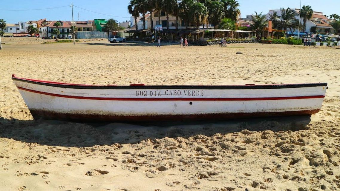 Una barca bianca con le parole 'Bom Dia Cabo Verde' sul fianco riposa su una spiaggia sabbiosa di fronte a una città costiera.