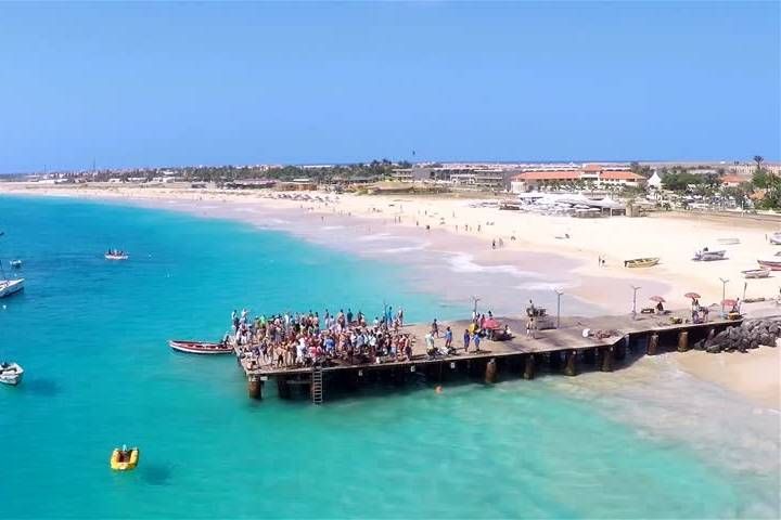 Un viaggio di gruppo WeRoad si riunisce su un molo di legno sull'acqua turchese, con una lunga spiaggia sabbiosa e una città costiera sullo sfondo.