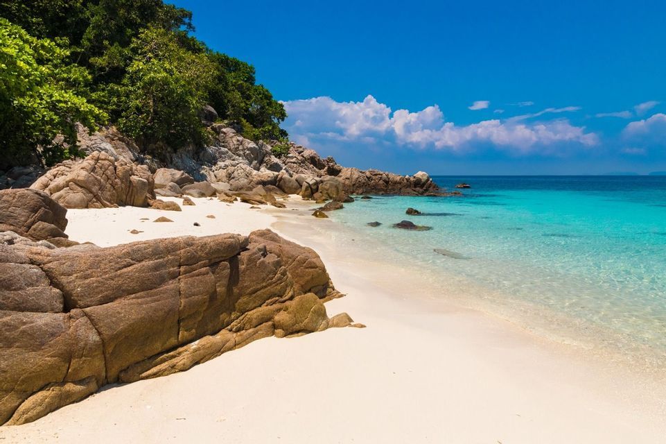 A secluded white sand beach with large rocks borders clear turquoise water under a blue sky.