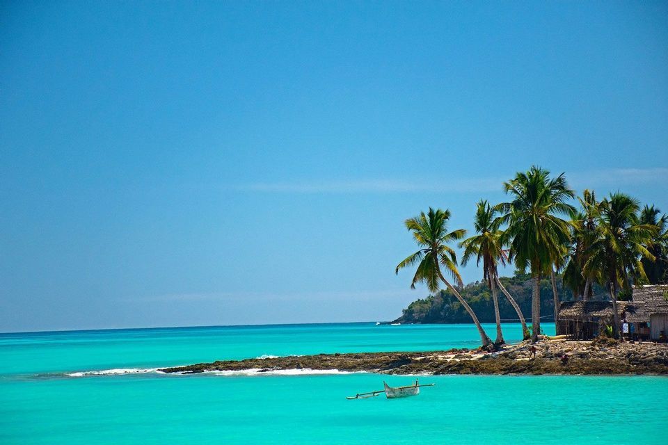 Una canoa de vela blanca flota en agua turquesa junto a una pequeña isla rocosa con palmeras y una cabaña bajo un cielo despejado.