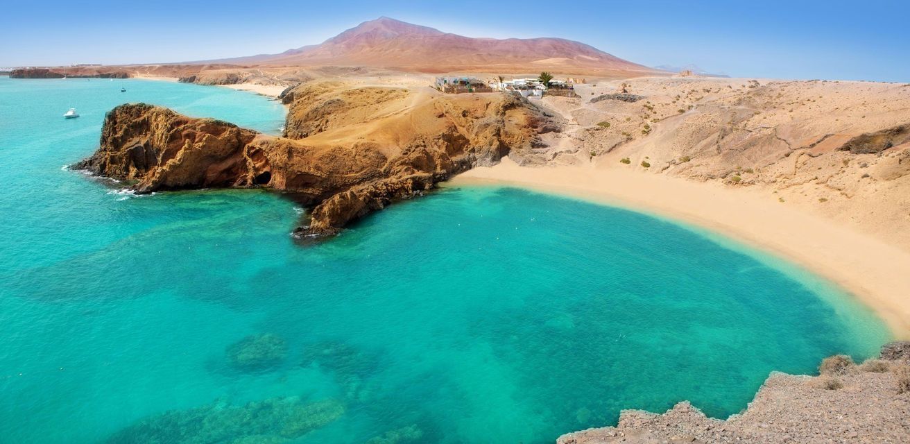 Una spiaggia sabbiosa si incurva attorno a una baia di acqua limpida e turchese, incastonata tra scogliere rocciose con una grande montagna in lontananza.