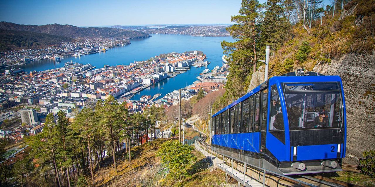 Una funicolare blu sale su un binario su un pendio soleggiato, offrendo una vista panoramica di una città costiera e del suo porto sottostante.