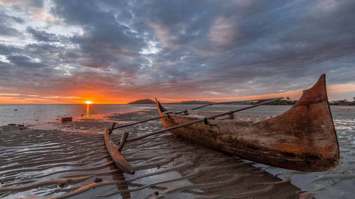 Una canoa a bilanciere in legno riposa su una spiaggia sabbiosa e umida con la bassa marea, durante un tramonto nuvoloso sull'oceano.