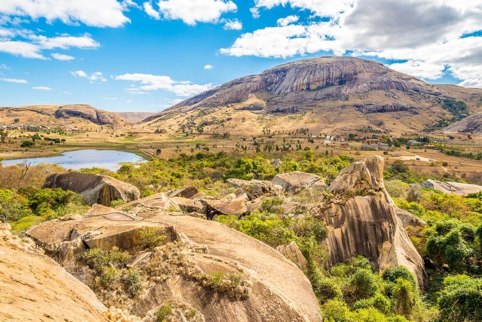 Un'ampia valle con un lago e grandi massi, che conduce a un'imponente montagna rocciosa sotto un cielo blu parzialmente nuvoloso.