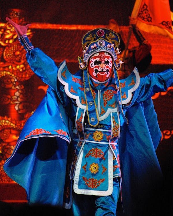 Un artista in costume tradizionale dell'opera cinese, con una tunica blu e una elaborata maschera rossa, posa sul palco.