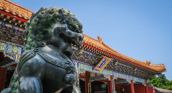 Una statua in bronzo di un leone guardiano cinese davanti a un tempio tradizionale con un tetto decorato di tegole arancioni sotto un cielo azzurro limpido.