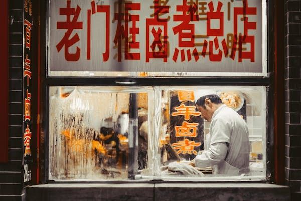 Un uomo in uniforme bianca che prepara cibo all'interno di un negozio, visto attraverso una finestra appannata sotto un'insegna con caratteri cinesi rossi.