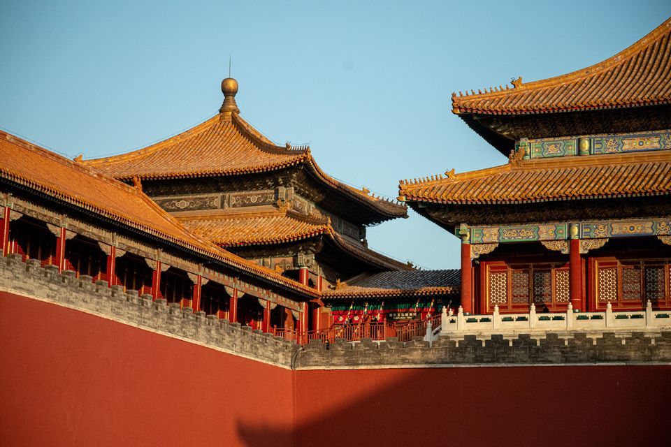 I tetti dalle tegole dorate degli edifici tradizionali cinesi si ergono sopra un muro rosso sotto un cielo azzurro chiaro.