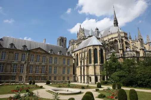 Un edificio classico e una cattedrale gotica visti da un giardino curato con aiuole e una piccola fontana sotto un cielo parzialmente nuvoloso.