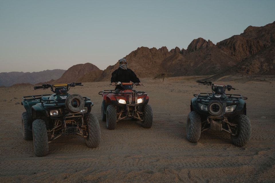 Una persona che indossa una kefiah è seduta su un quad rosso, affiancata da altri due quad, in un deserto con montagne sullo sfondo.
