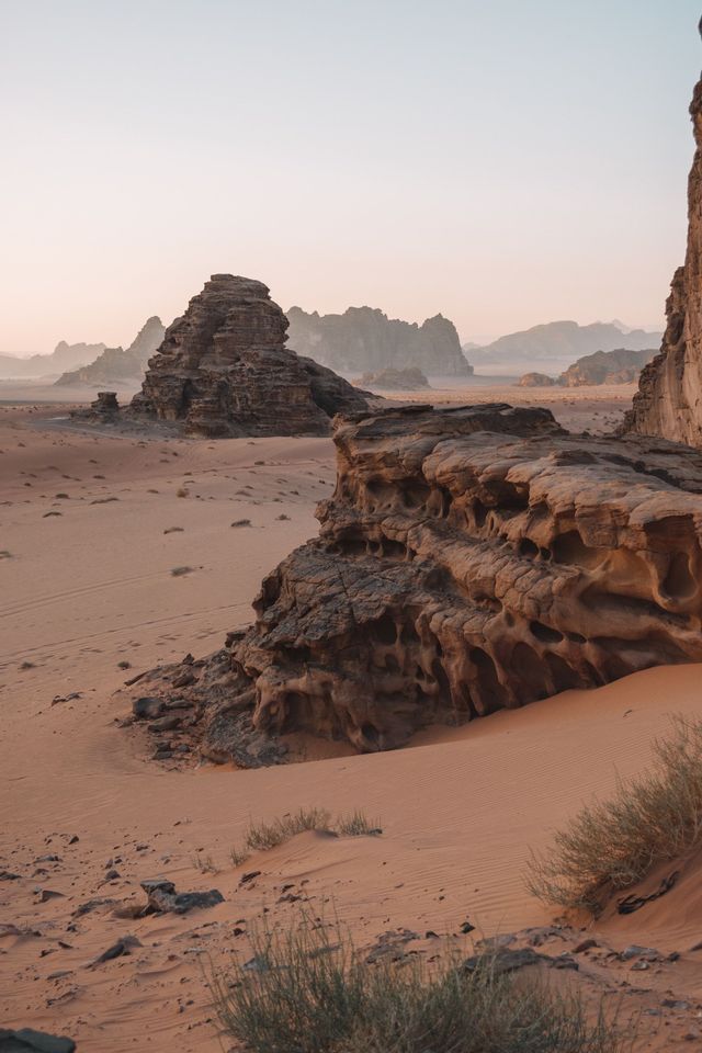 Grandi formazioni rocciose erose si ergono dalla sabbia rossa del deserto con arbusti sparsi, sotto un cielo pallido e fosco.